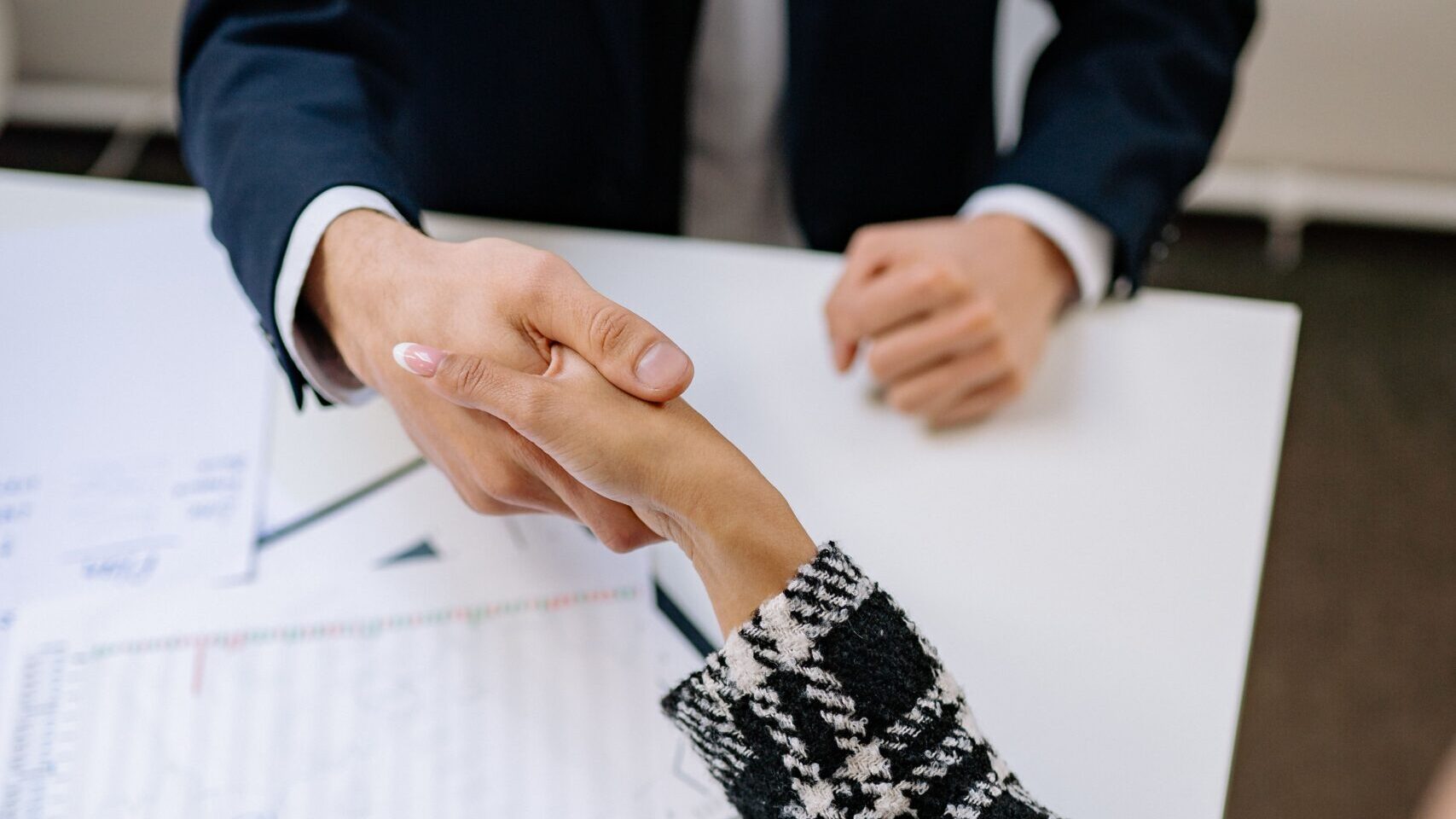 A close-up photo of a business handshake over financial documents in an office environment.